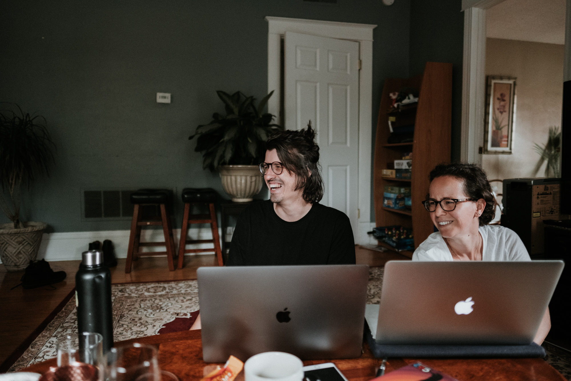 Two Modern Tribe employees sit at a table in a home working remotely on their laptops and smiling.